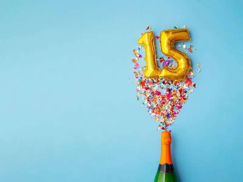 Golden number 15 balloons are bursting out from the top of a champagne bottle with a spray of colorful confetti against a serene blue background, conveying a festive and jubilant celebration of a 15-year anniversary.