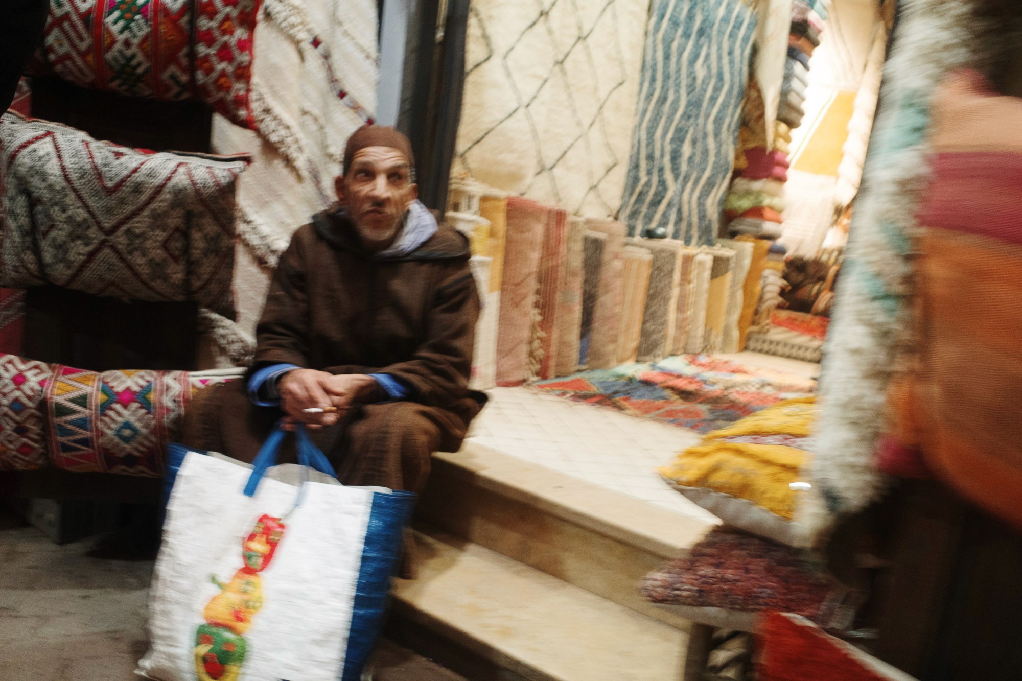 A person in a brown hooded garment sits in a marketplace surrounded by colorful, intricately patterned textiles and cushions, holding a white bag with a colorful design.