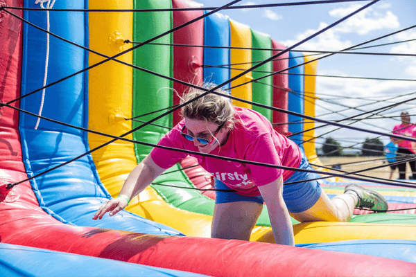 Outdoor team-building event with participants wearing matching blue shirts, taking part in inflatable obstacle courses and games, smiling and cheering, with the words 'ENTER A TEAM' in large bold white and green text across the image.