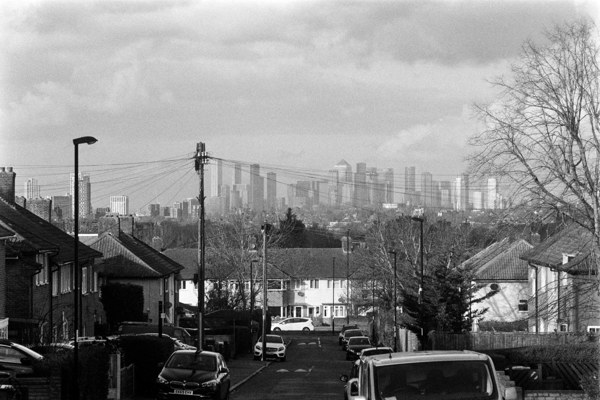 Residential street view looking toward distant City of London skyline, semi-detached houses with parked cars, overhead telegraph pole with radiating cables in mid-ground
