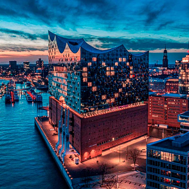 A green suspension bridge spanning across a body of water, with forested hills and residential homes in the background.