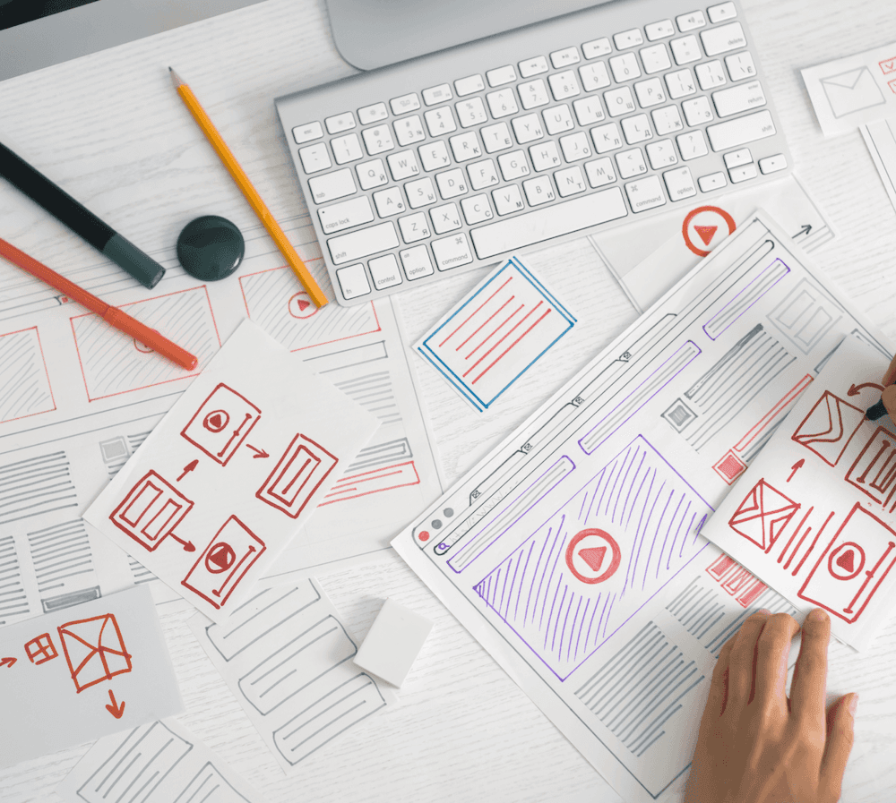 Schreibtisch mit Tastatur, Stiften und mehreren Papierentwürfen für Webseitendesign, eine Hand hält einen Entwurf mit Symbolen und Textblöcken