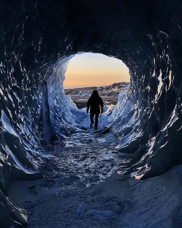 A person walking out of a blue ice cave toward the sunset