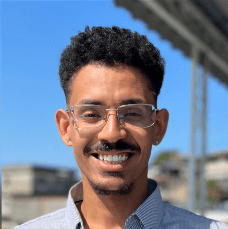 A man with curly hair, glasses, and a friendly smile, standing outdoors with a blue sky in the background.