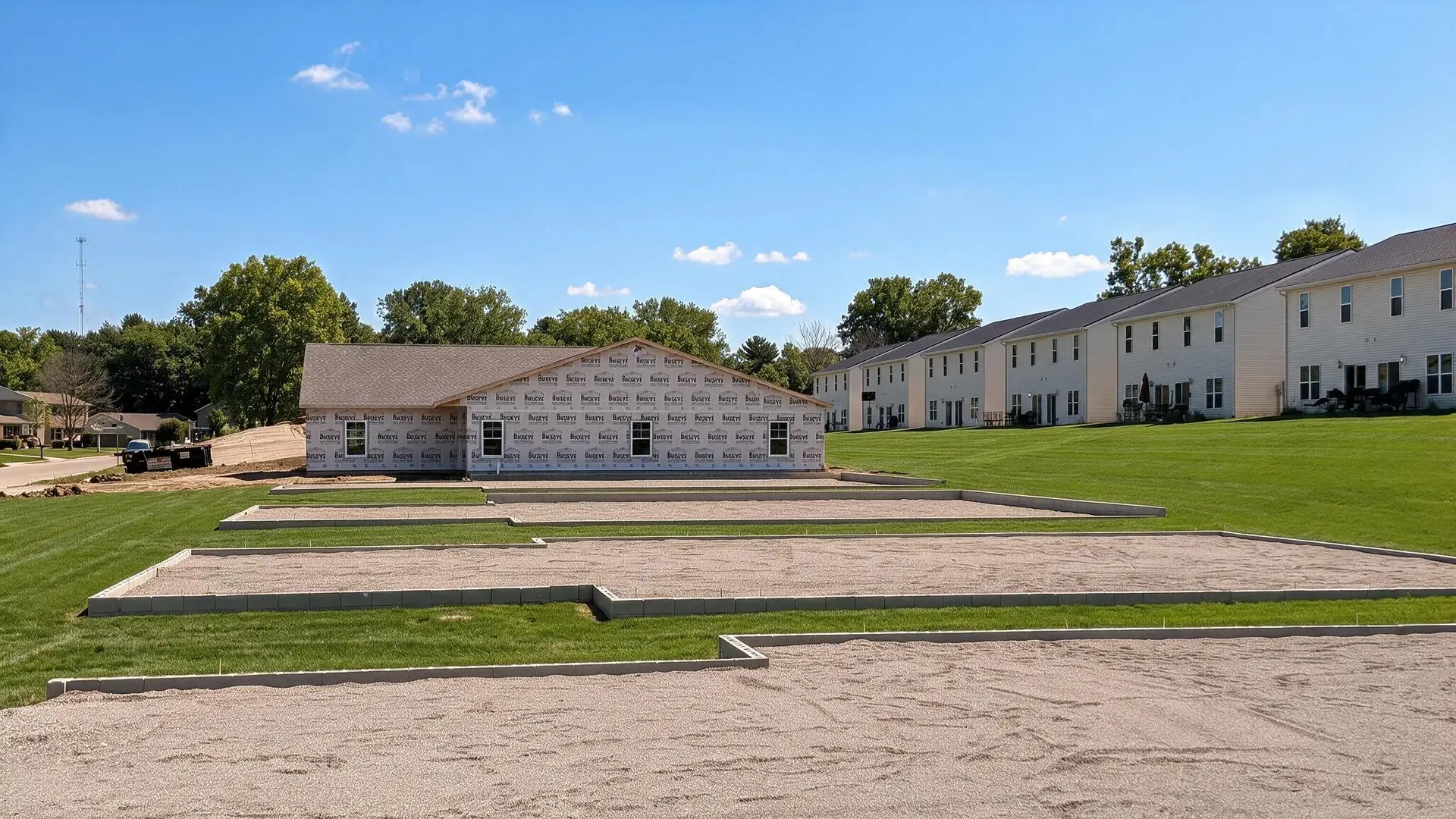 Freshly poured concrete foundations laid out on a cleared lot, with a wrapped house behind and existing apartments to the right