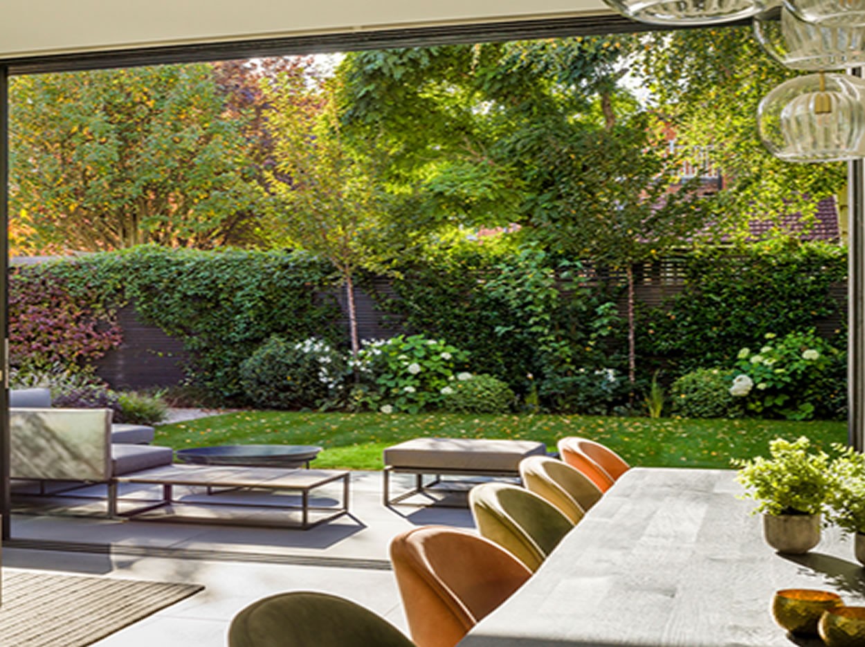 A modern outdoor dining area opens to a lush garden, featuring a table, chairs, and vibrant greenery in the background.