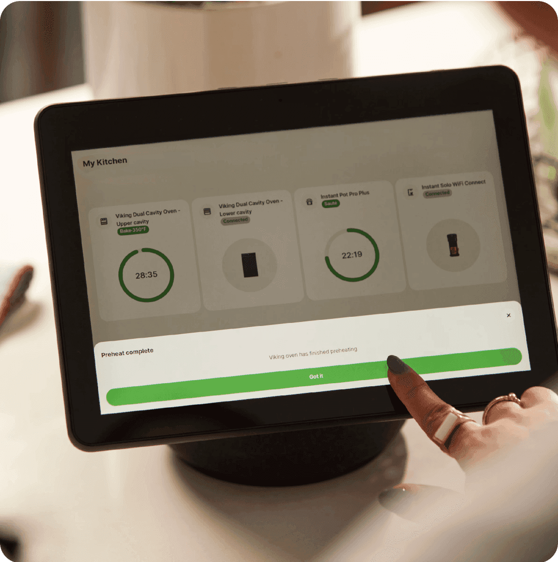 ablet displays "My Kitchen" dashboard with multiple connected appliances showing timers like "28:35" and "22:19" while a user taps a green status bar reading "Preheat complete"