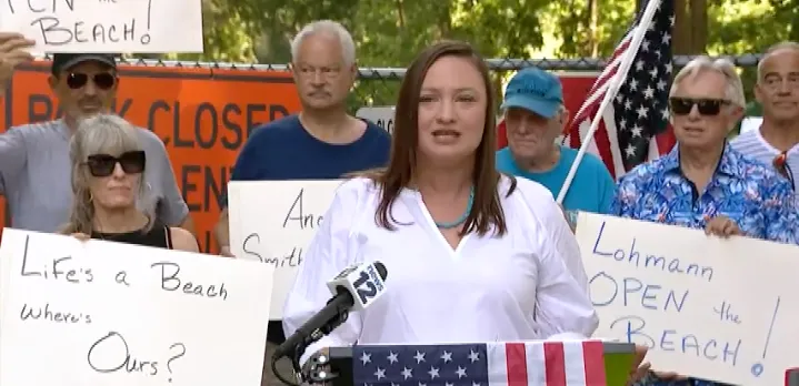 A woman speaks at a rally, surrounded by supporters holding signs, with trees in the background.