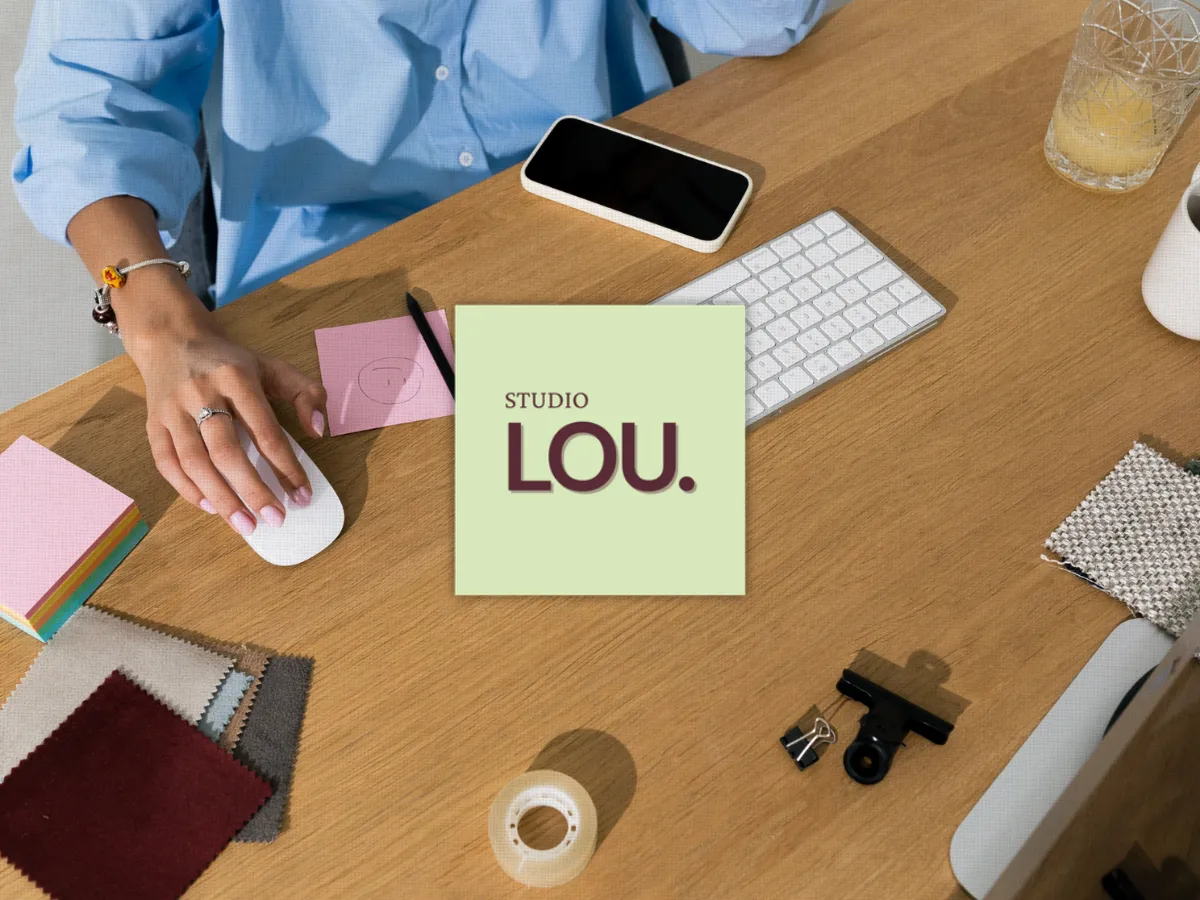 Top-down view of a person working at a wooden desk with a computer keyboard, mouse, smartphone, and design swatches, overlaid with a green graphic reading "Studio Lou."
