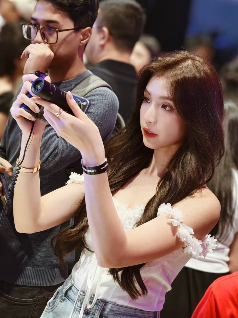 A young woman with long hair holds a camera, focused on capturing an image, while a man in the background appears to be deep in thought.