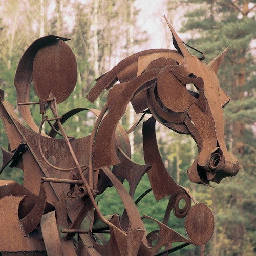 Rusty metal sculpture of a horse's head, set against a background of trees.