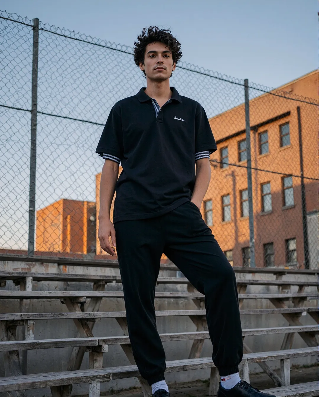 Fashion portrait featuring black polo and pants against urban backdrop with brick buildings and stadium seating during golden hour