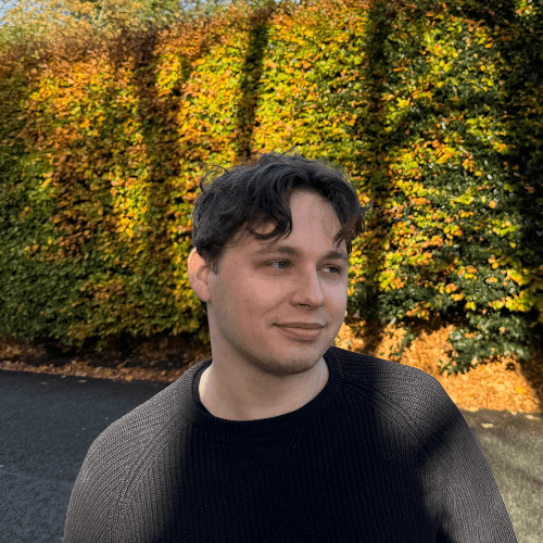 Person wearing a blue puffer jacket and red backpack standing in front of a light blue building on a city street.