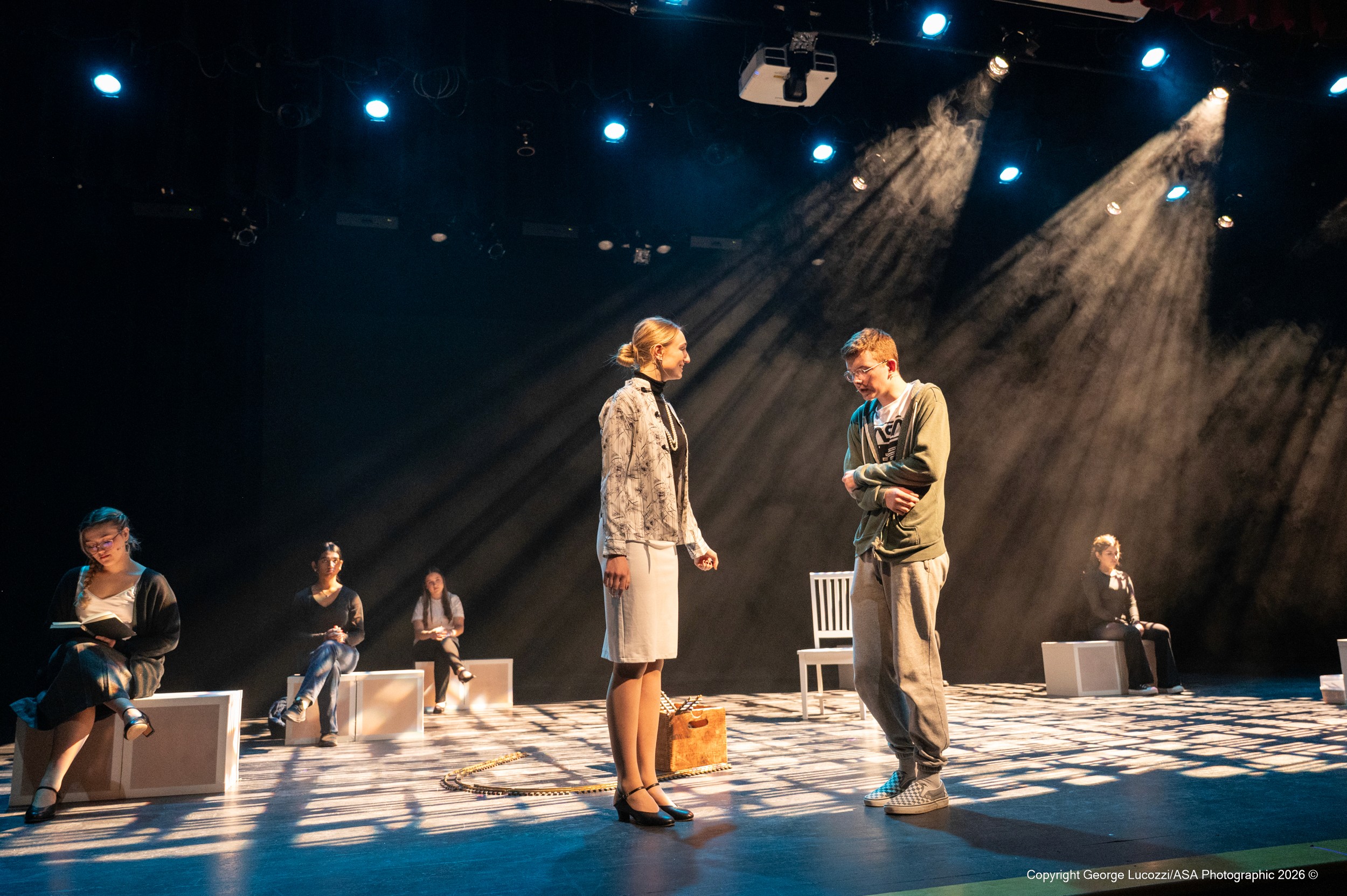 Downstage, Ms. Alexander and Christopher stand in intense, sunlike tungsten beams that cut visibly through heavy haze, while the “Voices” sit casually on cubes upstage, evoking a park atmosphere in softer spill light.