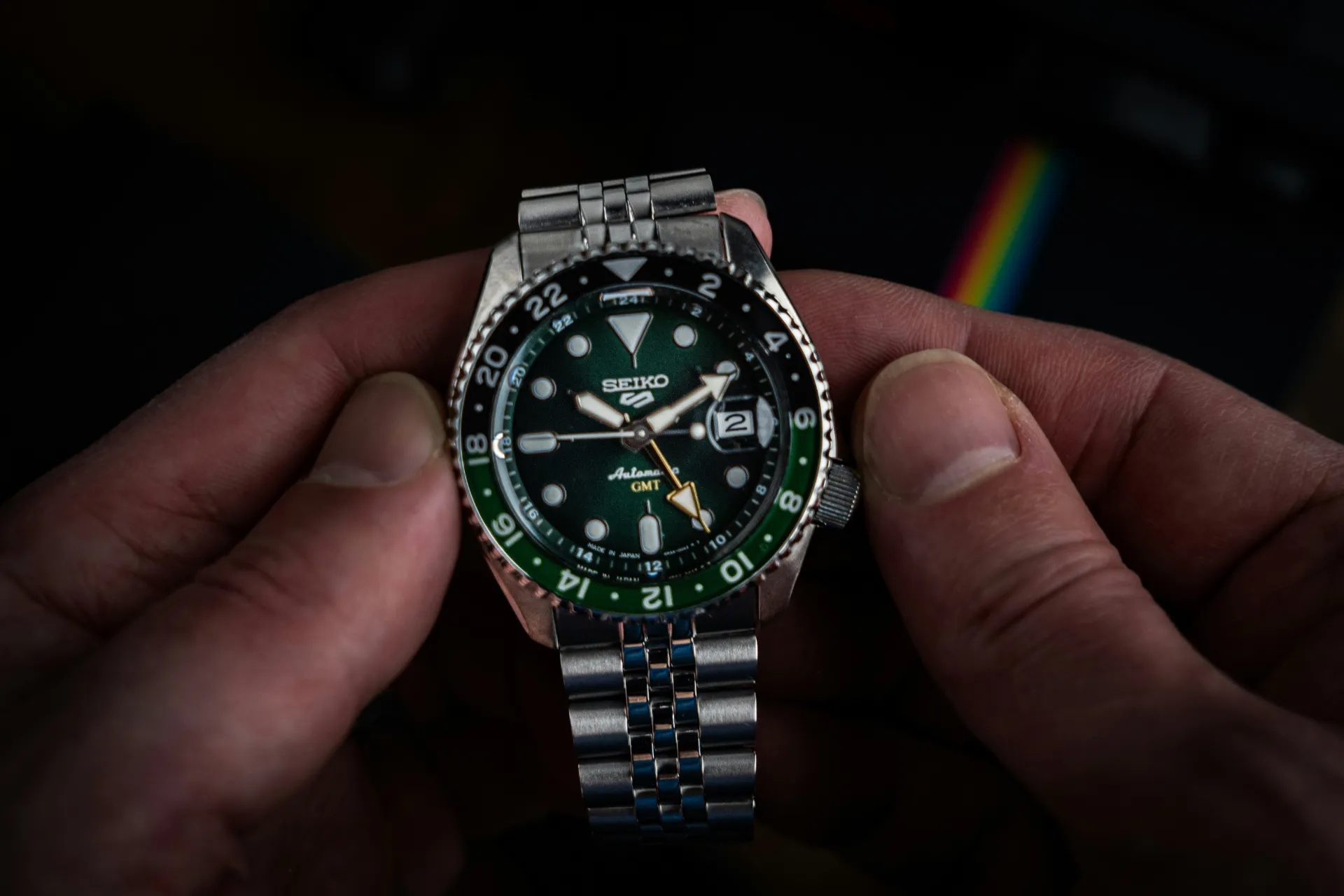 Seiko 5 Sports automatic GMT wristwatch with green sunburst dial, green 24-hour bezel, and stainless steel bracelet, held between two hands against a dark background.