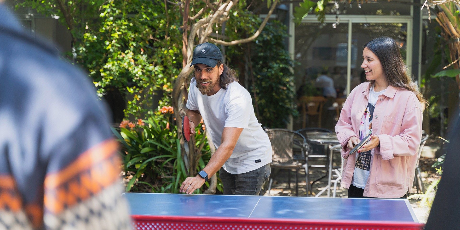 Dos personas jugando tenis de mesa mientras otra los observa. De fondo se ven árboles