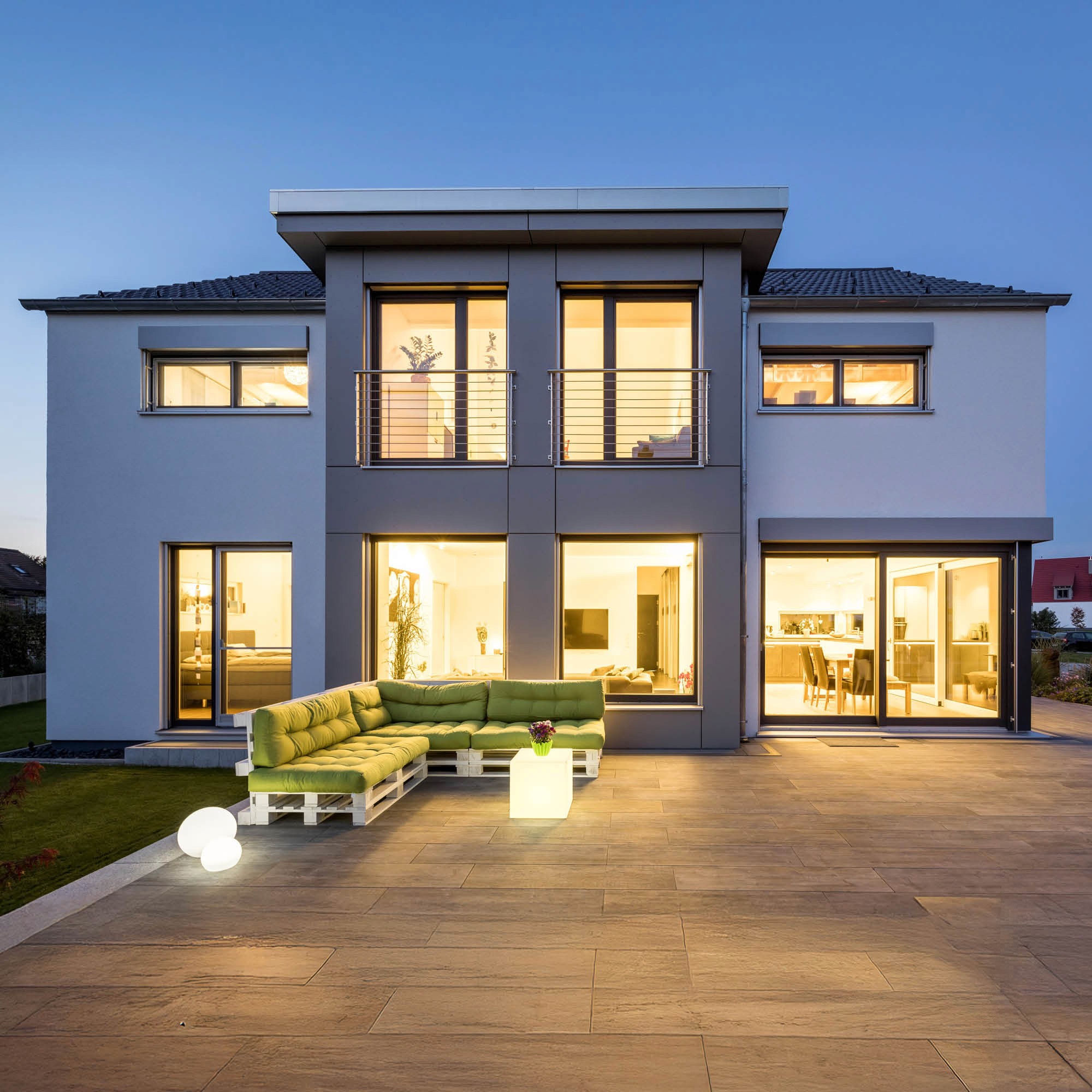 Moderne zweistöckige weiße Villa mit großem Fenster, Balkon, heller Lounge-Terrasse mit Sofa und beleuchteten Kugeln.