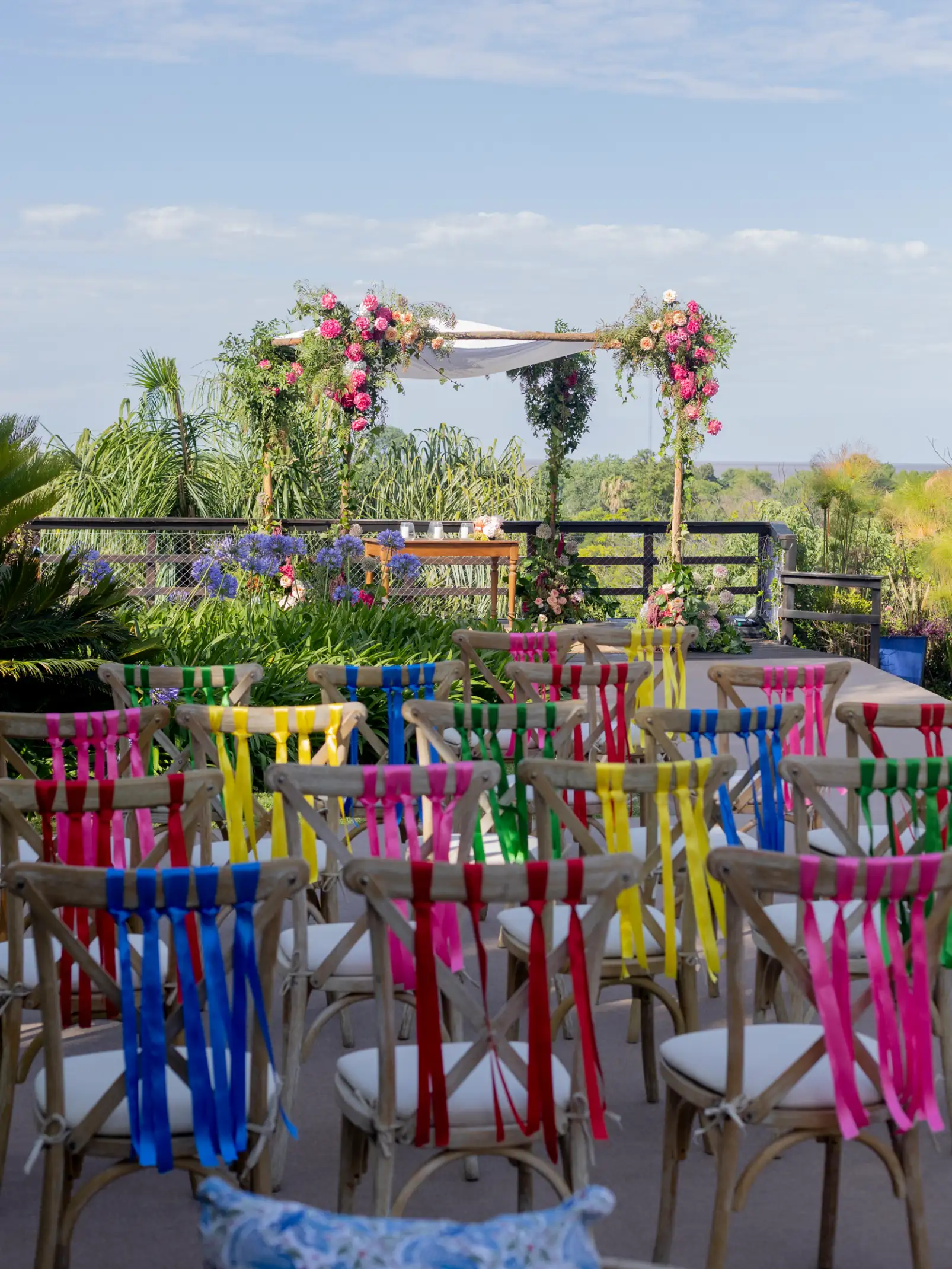 Escenario de ceremonia al aire libre con sillas coloridas y arco floral para evento festivo, evocando alegría y naturaleza tropical.