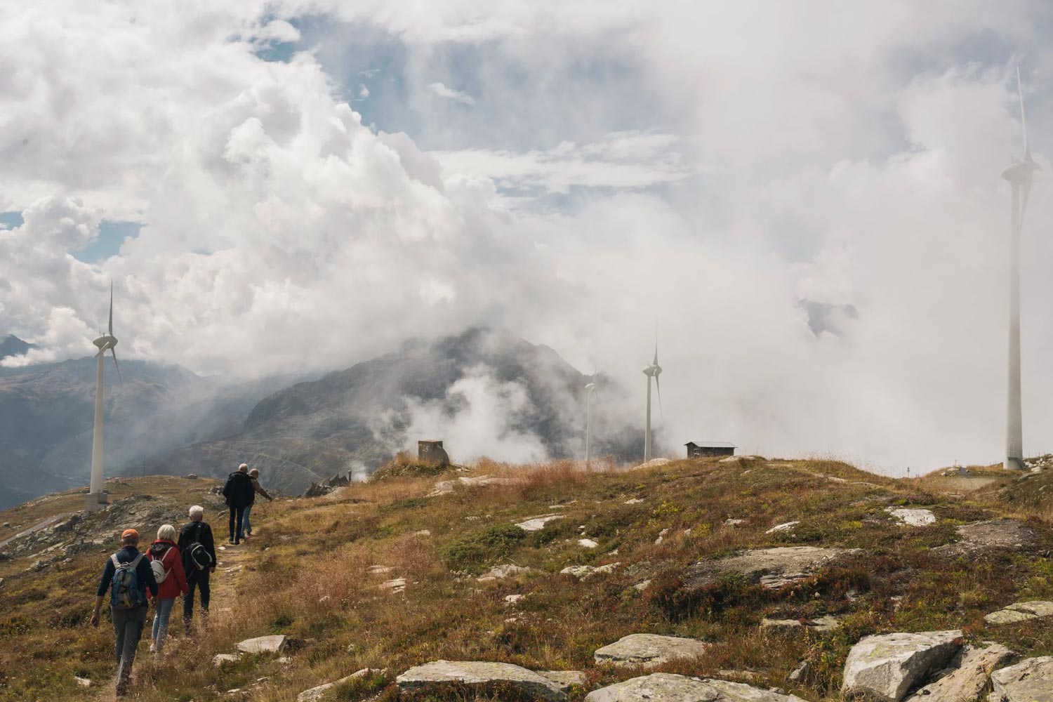 Das Foto zeigt mehrere Wanderer in einer weitläufigen, alpinen Gebirgslandschaft. Im Vordergrund spazieren Personen über einen grasbewachsenen, von Steinen durchsetzten Bergpfad. Eine Frau in der linken Dreiergruppe hält an, um mit ihrem Smartphone die Aussicht zu fotografieren. Auf der rechten Bildhälfte ragt eine große, moderne weiße Windkraftanlage markant in den Himmel. Links unten, tiefer am Hang, ist eine dunkle Holz-Berghütte zu erkennen. Im Hintergrund erstrecken sich massive, tiefe Täler und hohe Bergketten, deren Gipfel teilweise in dichten, tiefhängenden weißen Wolken verschwinden. Die Szenerie vermittelt eine dramatische, aber friedliche Naturatmosphäre.