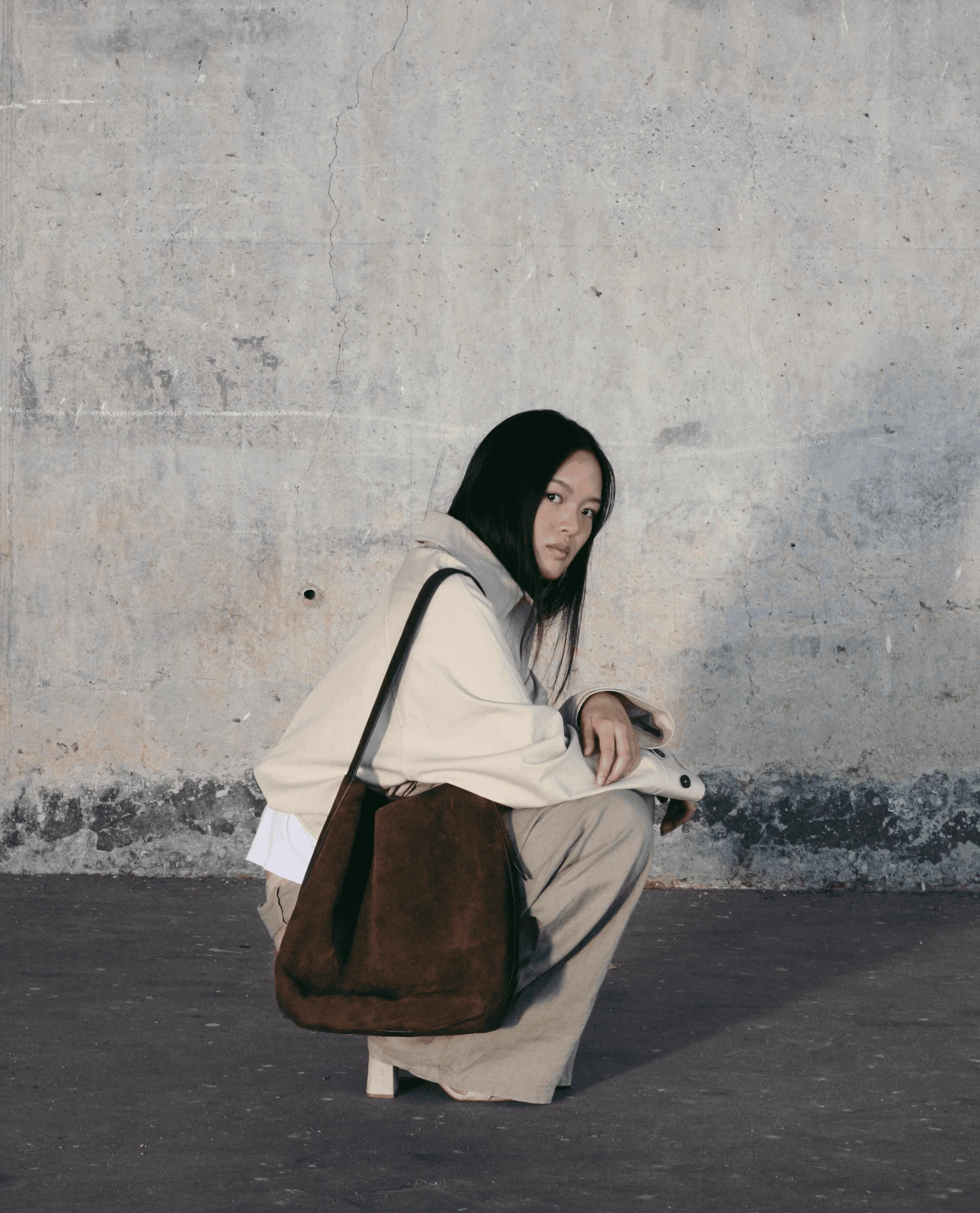 Modelo agachada posando en una pared brutalista con un bolso de la marca de bolsos artesanales Lulia Spain