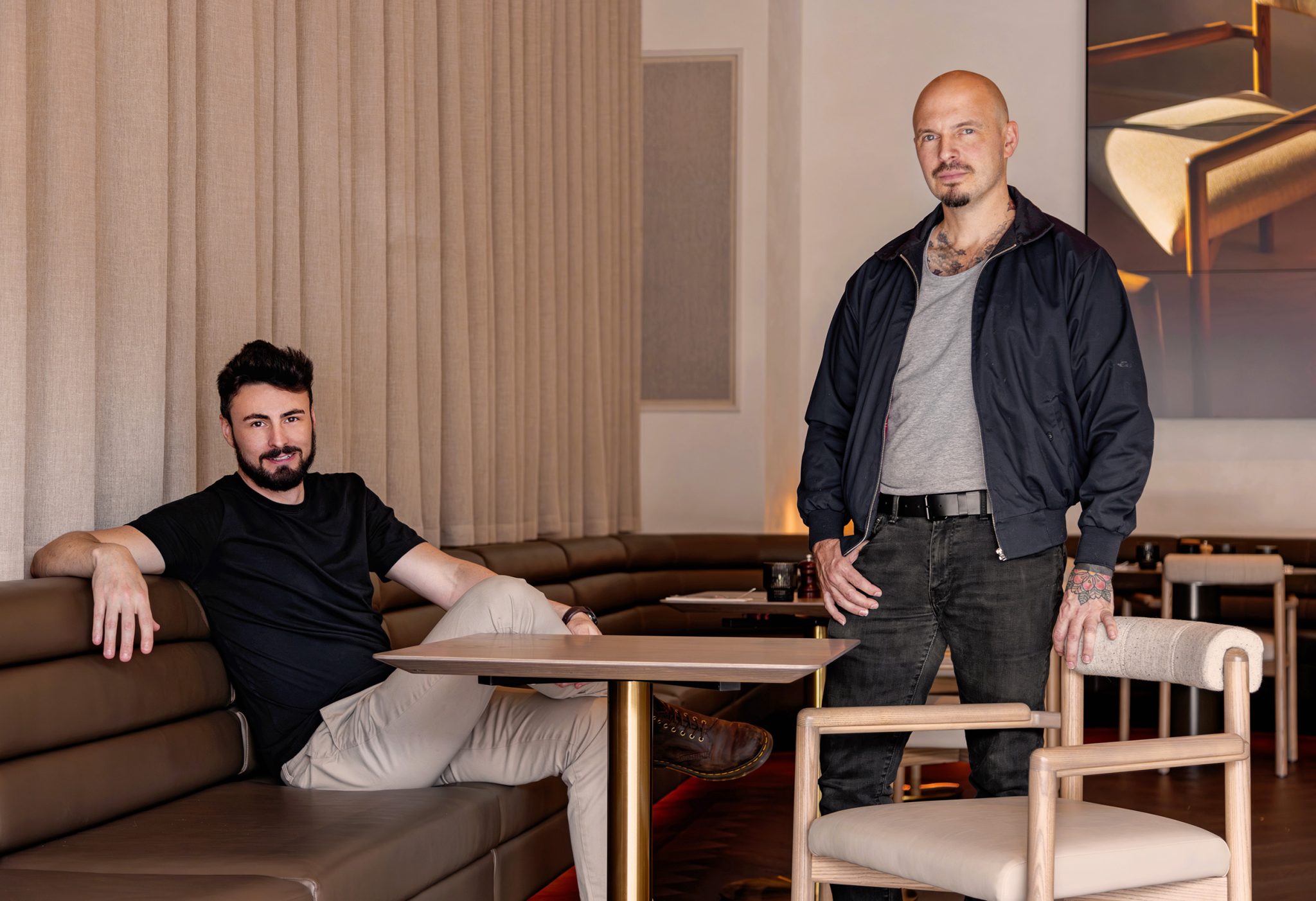 Two men, one seated and one standing in a bomber jacket with visible tattoos, in a lounge area with dining chairs