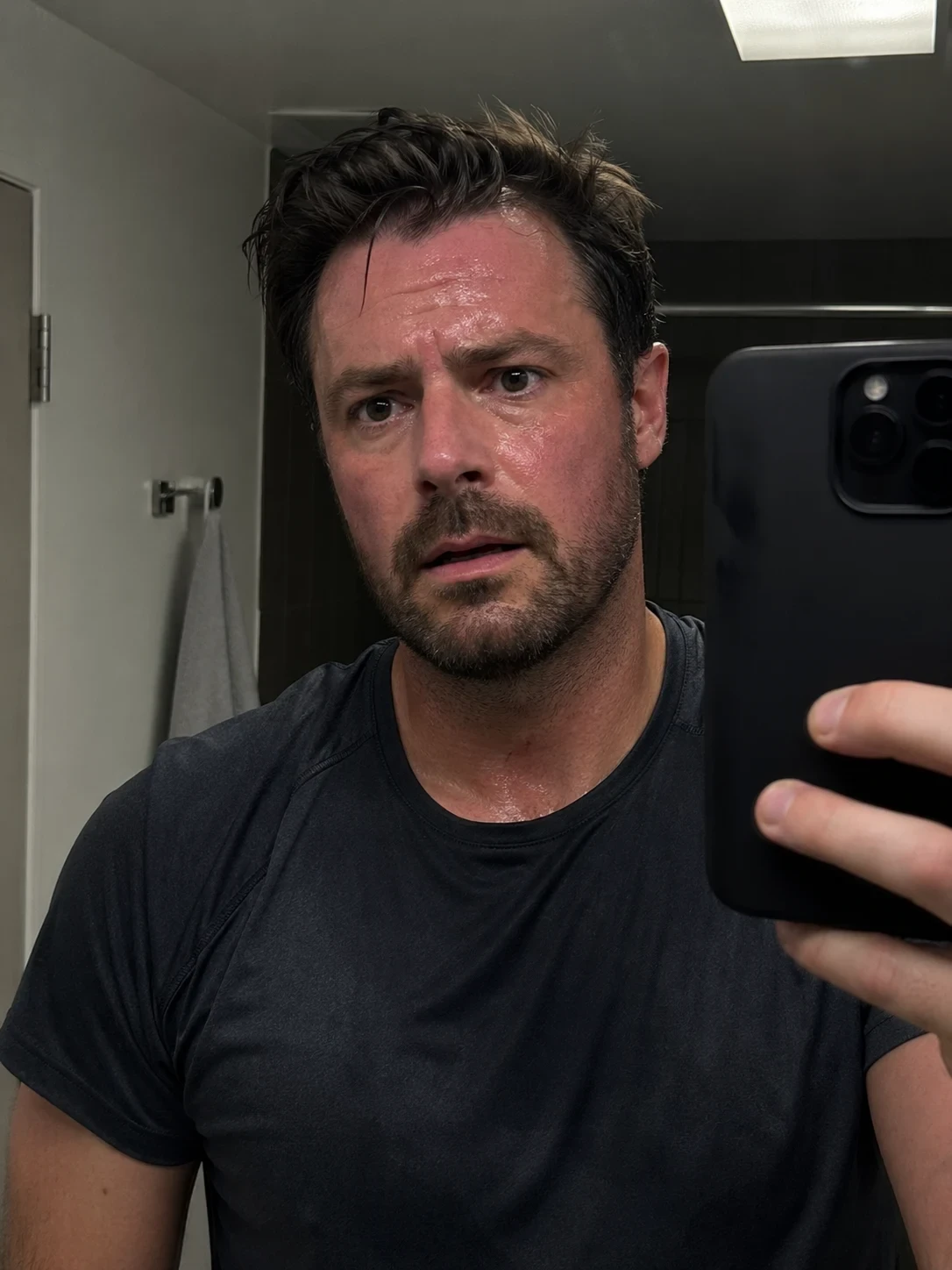 Sweaty post-workout bathroom mirror selfie with a tired expression and dark athletic shirt.