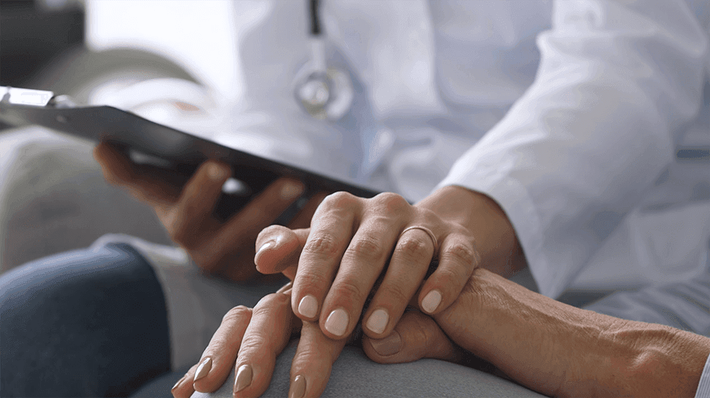 Close-up of doctor's hand comfortingly touching patient's hand while holding tablet, showing compassionate healthcare interaction.
