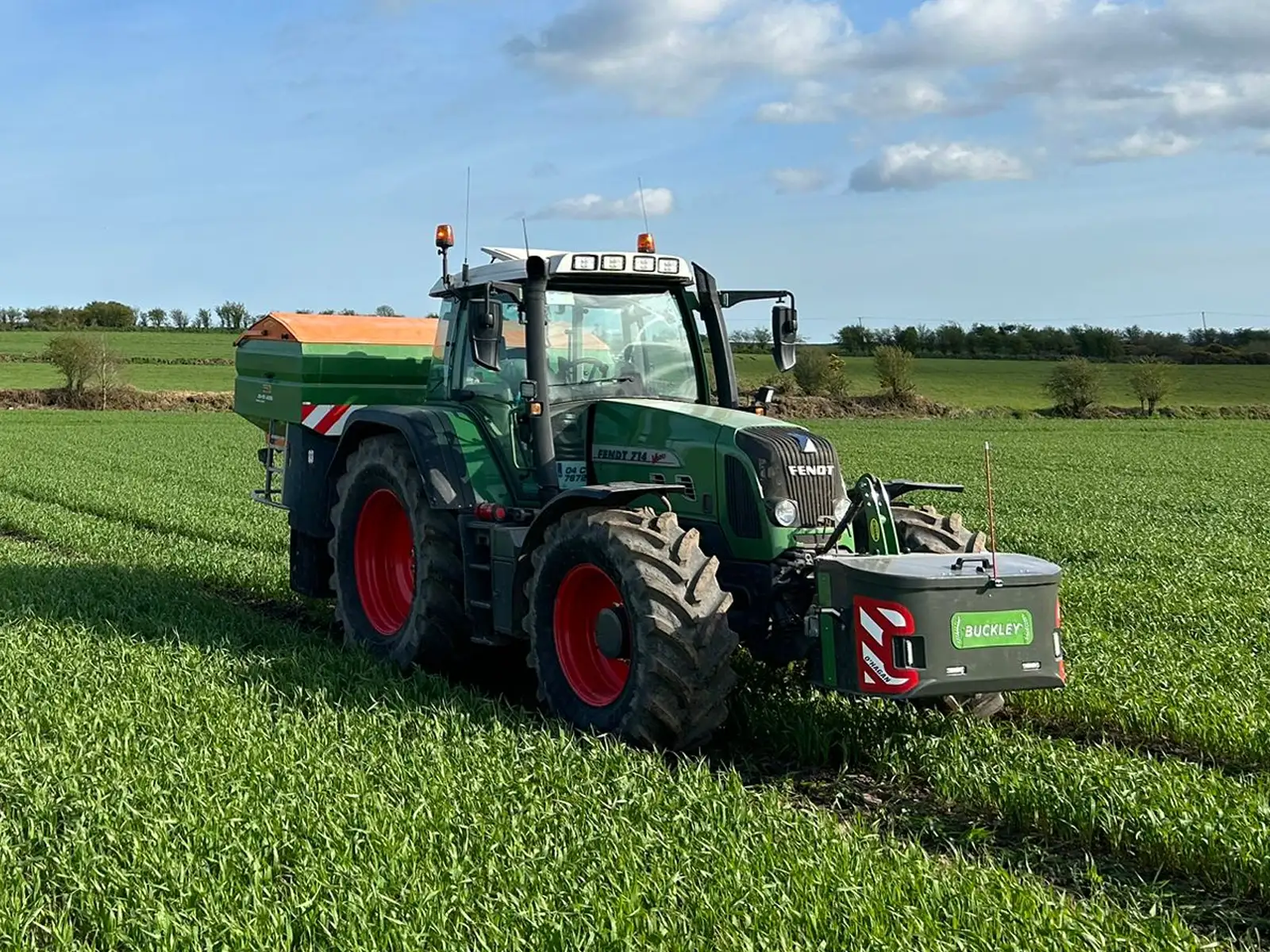 John Deere Green Tractor with black and yellow wheels on a country road with a green hydraulic bale handler attached beside an old wall with big trees in the background