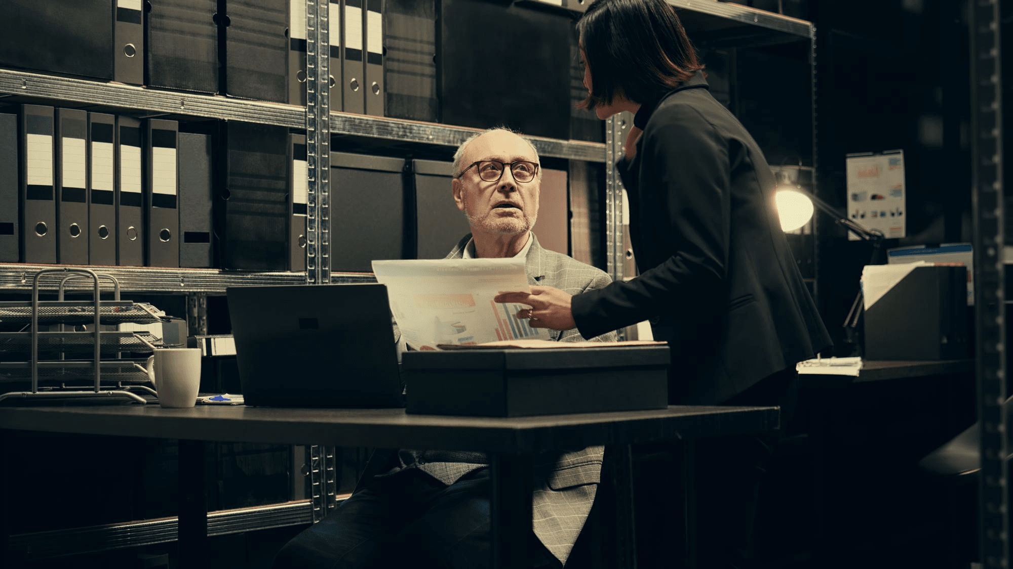 Two colleagues discussing printed charts in an office with shelves of binders in the background.