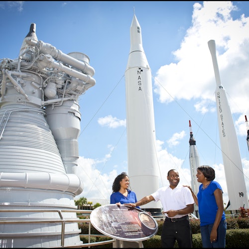 Tre personer står nära en NASA-raketutställning, med raketer i bakgrunden och en informativ skylt i förgrunden.