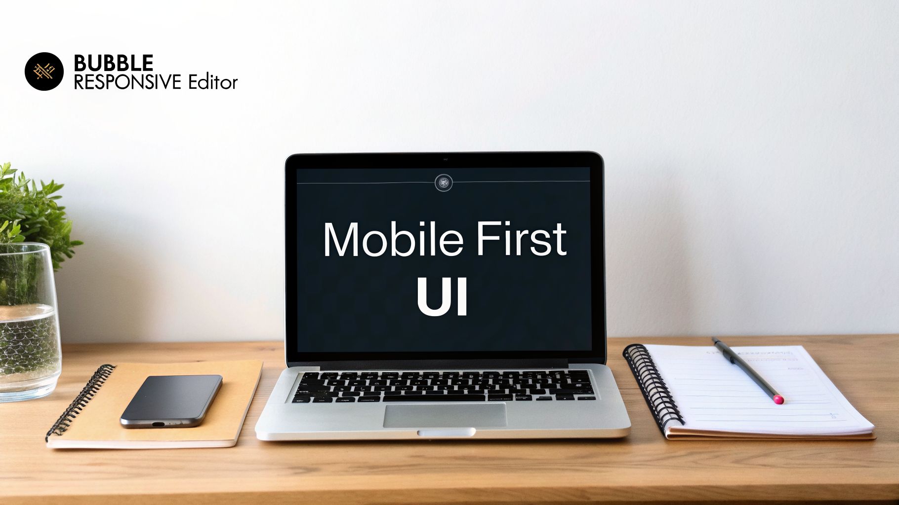 A laptop displaying 'Mobile First UI' on a wooden desk with a smartphone, notebooks, and a glass of water.