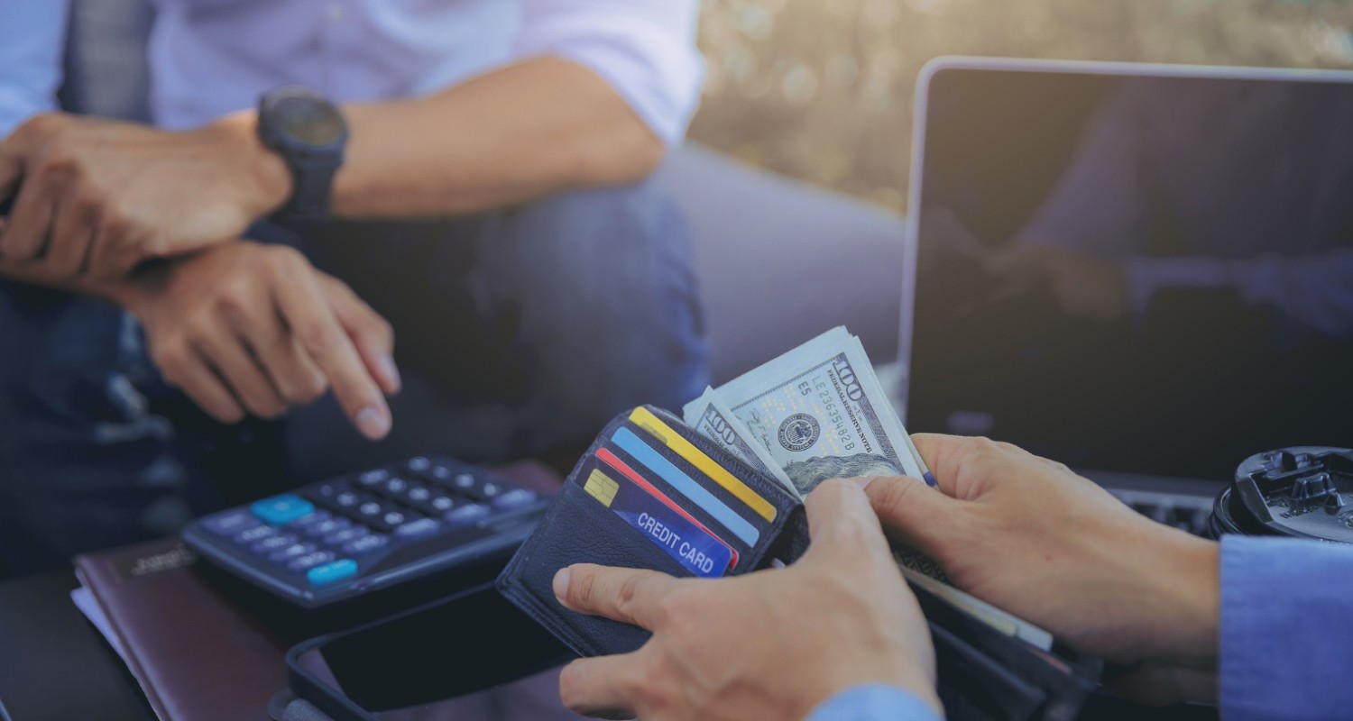 A person holds a wallet filled with credit cards and US dollar bills while another individual points at financial documents and a calculator, illustrating the integration stage of money laundering where illicit funds enter the legitimate economy.