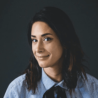 Smiling woman with dark hair wearing a checkered shirt and tie against a dark background. Her expression is warm and confident.