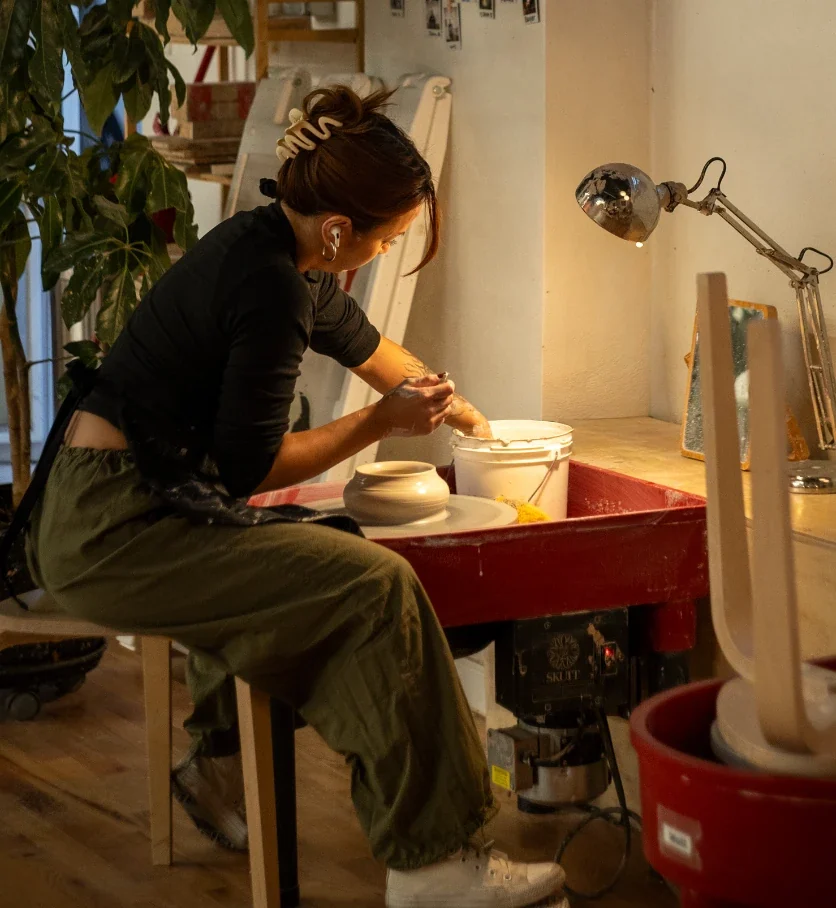 Member throwing a pot on a red Skutt wheel under a warm desk lamp at Kinship Studio