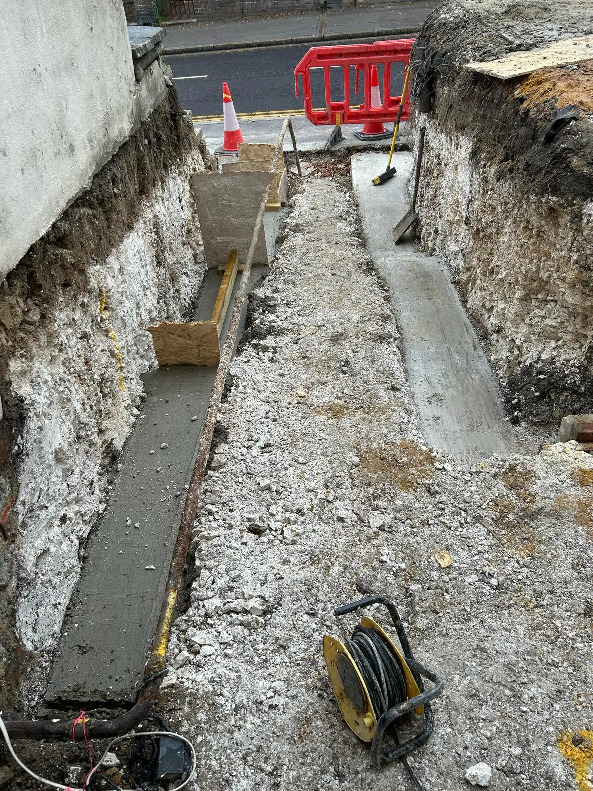 A narrow excavation site with gravel, tools, and a warning sign visible in the background.