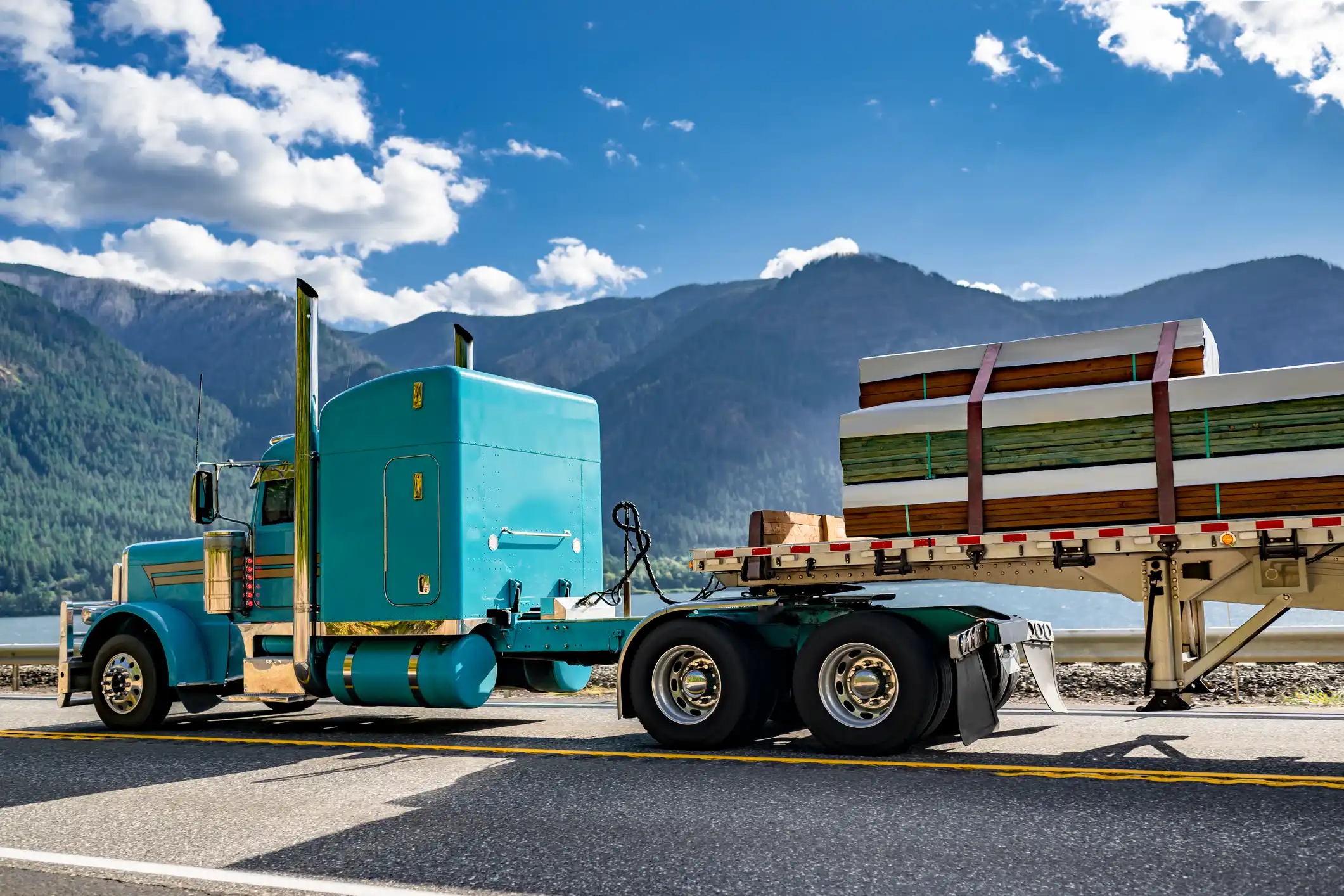 A teal semi-truck with a flatbed trailer hauling secured lumber along a scenic mountain highway — symbolizing GIA Group, LLC’s dependable support for truckers navigating carrier or coverage questions.