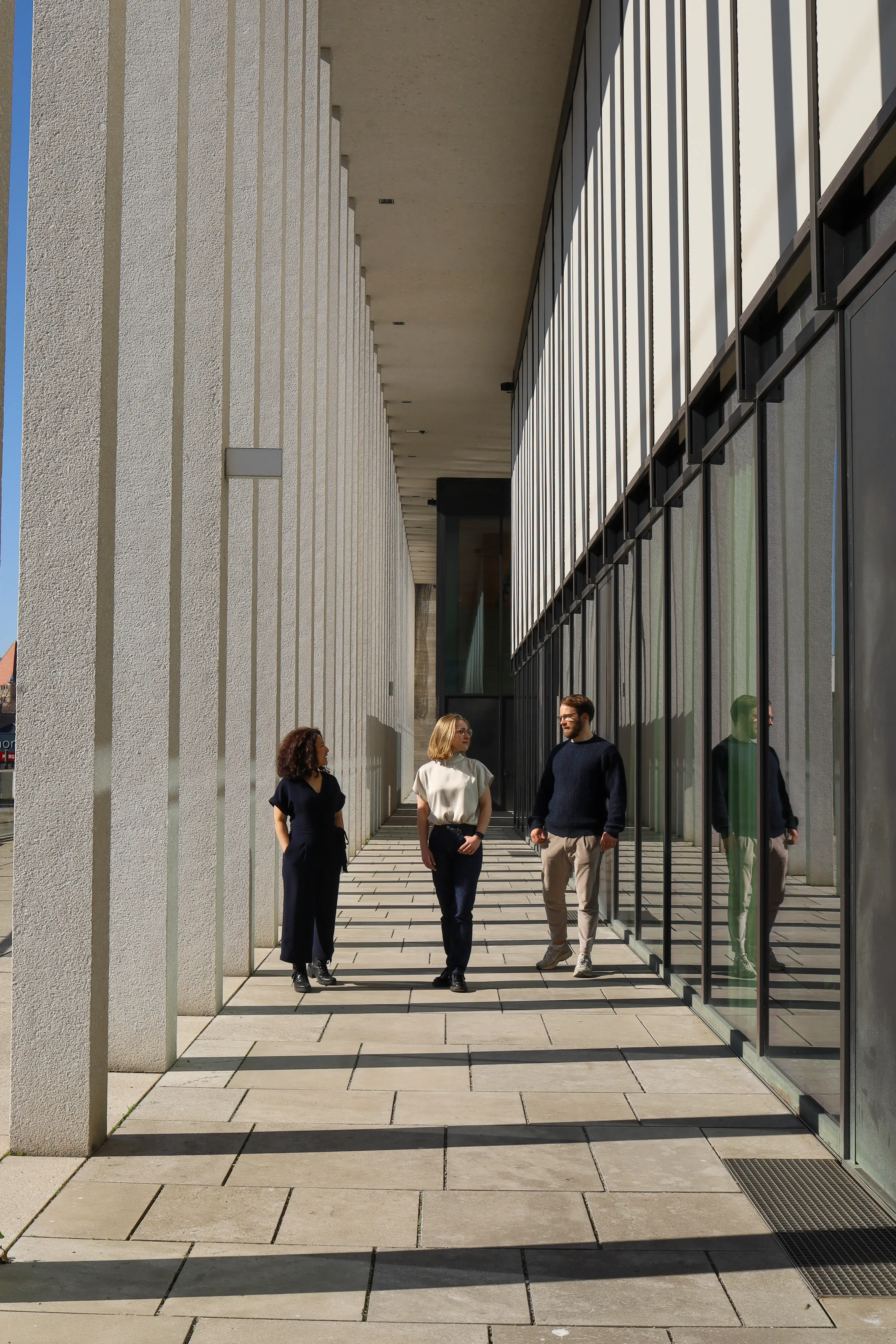 Jenia walking and chatting with professional colleagues in a modern architectural corridor.
