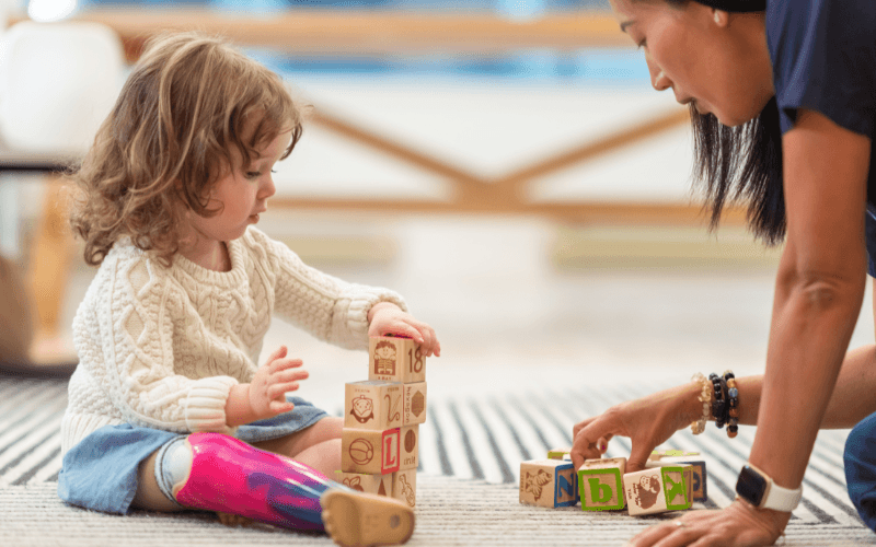 small child sits on rug in bright indoor room playing with blocks assisted by woman wearing navy blue scrubs