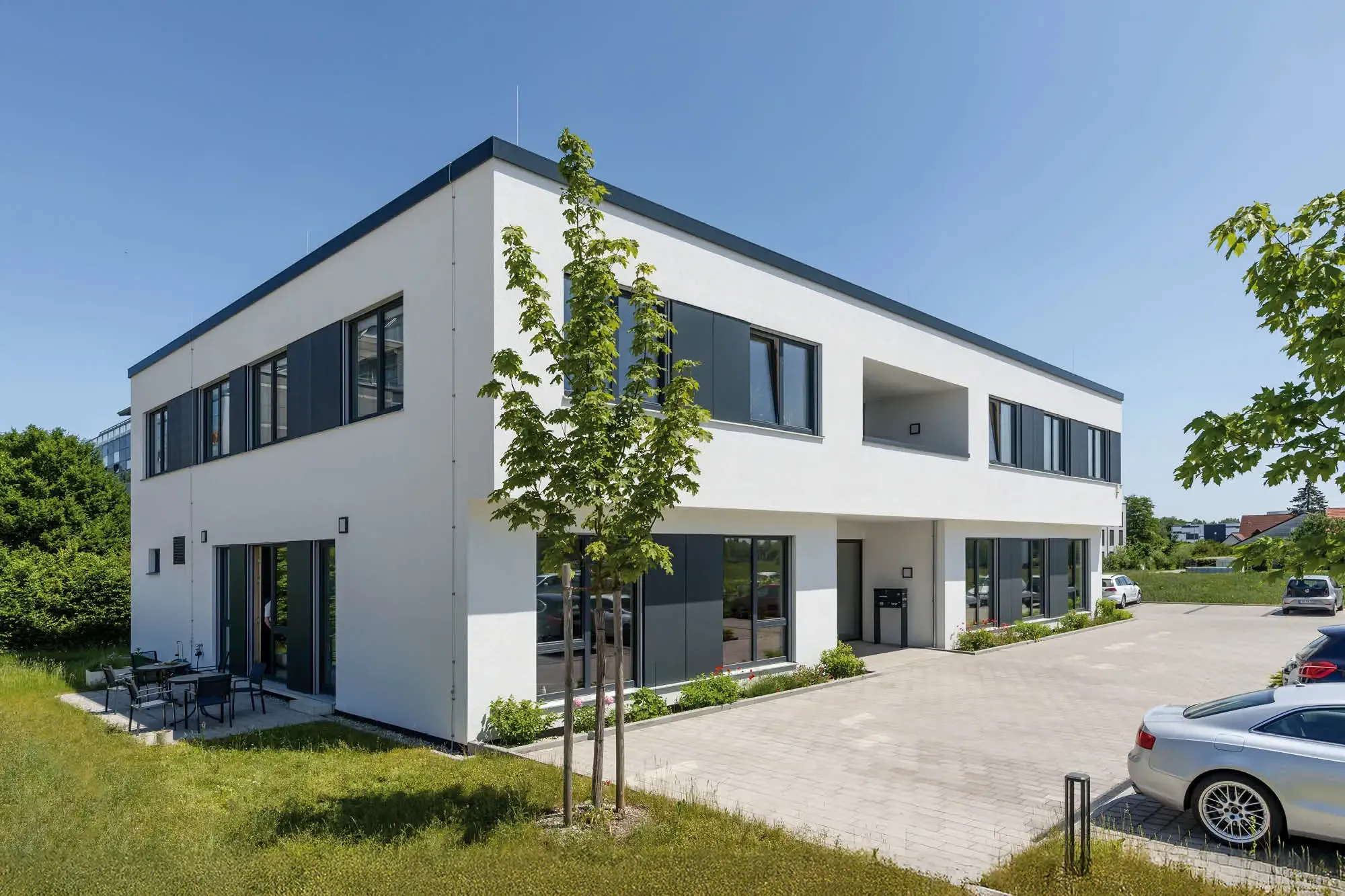 Modernes weißes Gebäude mit dunklen Fenstern und einem Flachdach vor blauem Himmel, mit Bäumen und einem Parkplatz.
