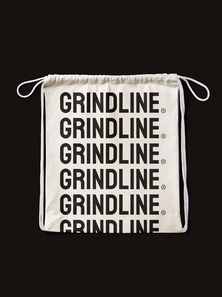Cream drawstring tote bag on black, printed front and centre with six stacked rows of the black “GRINDLINE®” wordmark.