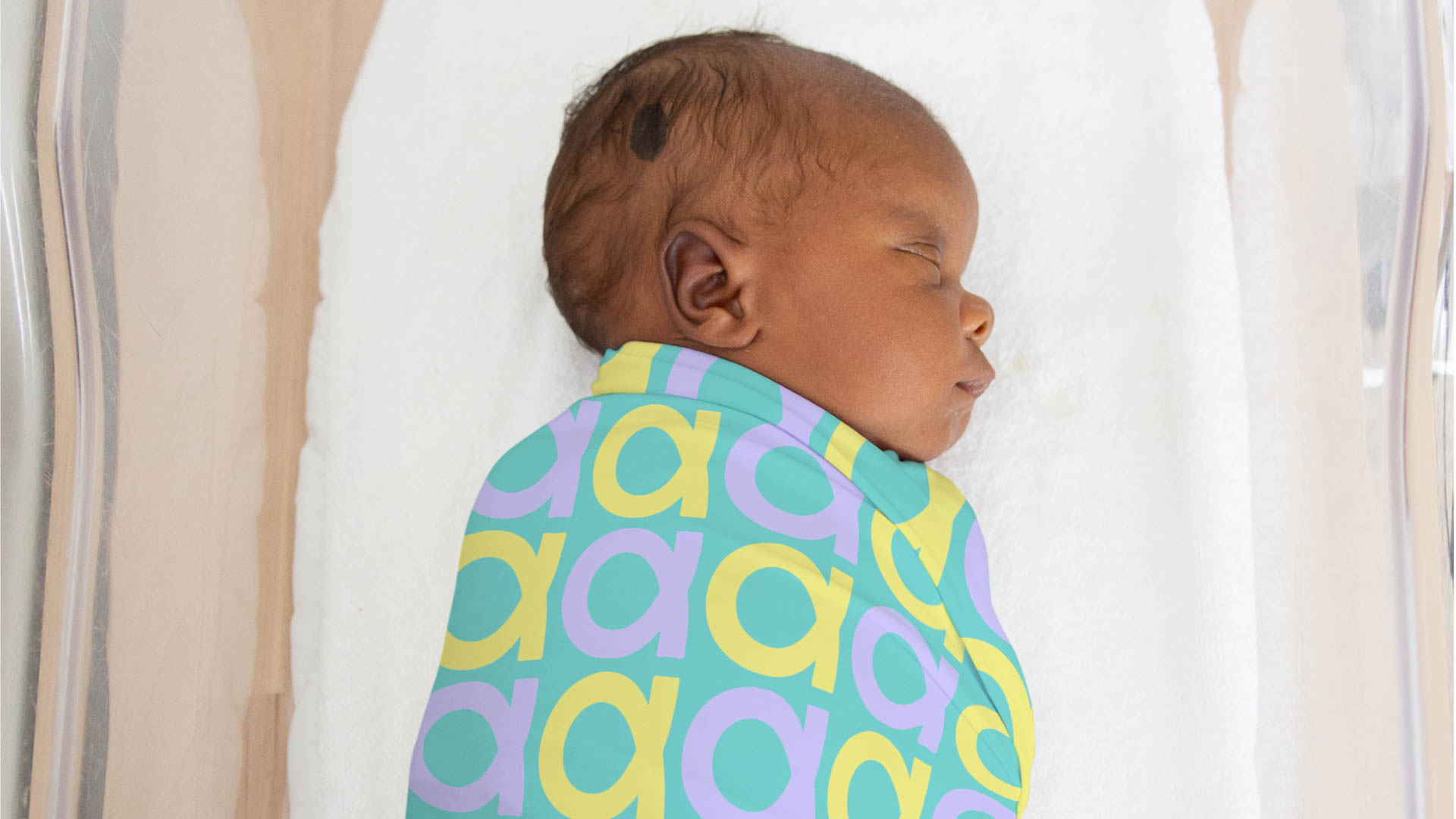 A newborn baby with dark skin and short hair is sleeping on a white surface. The baby is wearing a teal garment with a pattern of yellow, purple, and light blue letters. The garment covers the baby’s torso and shoulders. The baby’s head is turned to the right, and its eyes are closed. The background is a plain white surface.