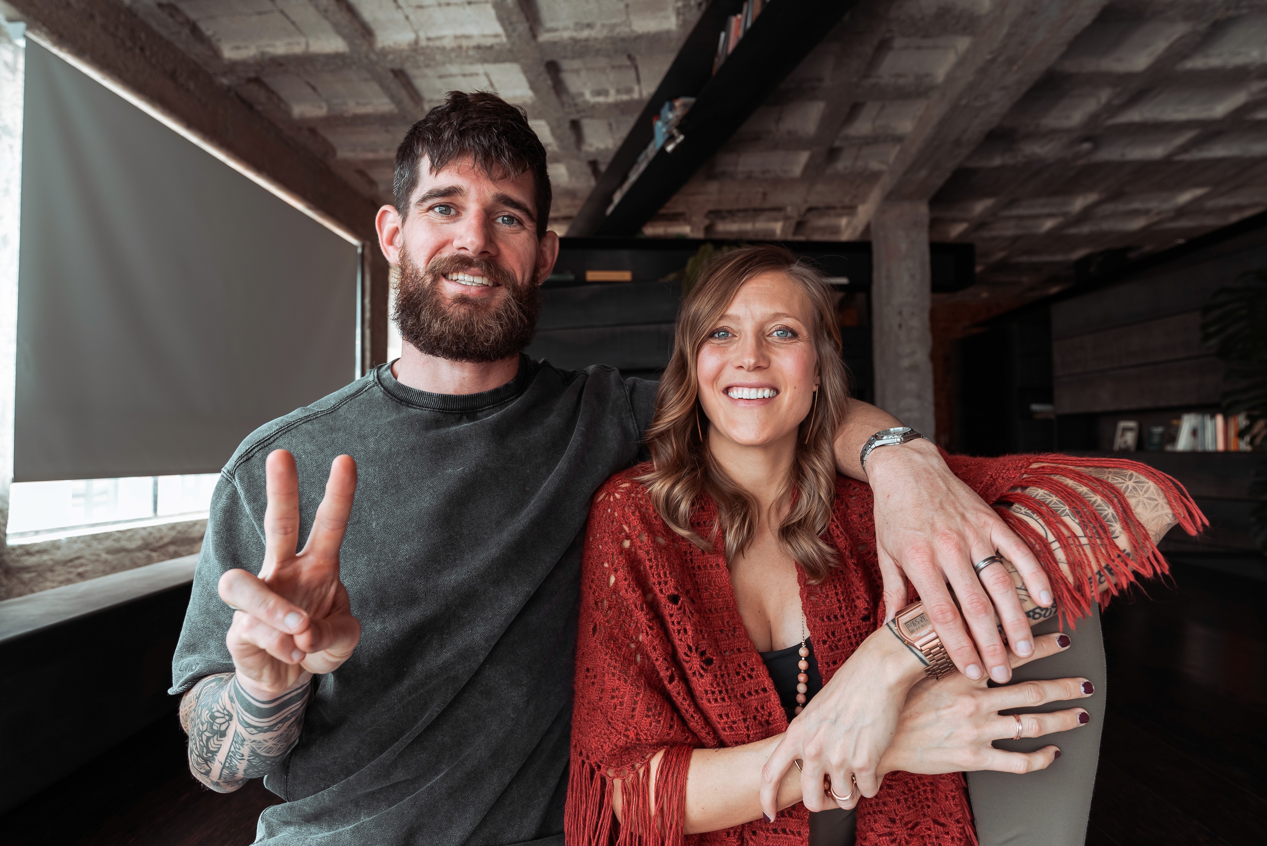 Bre and Flo smile towards the camera in a cozy room. He shows a peace sign, and she wears a red shawl.