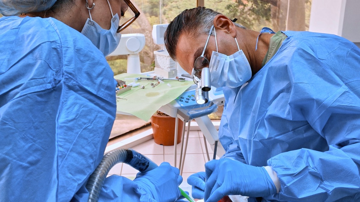 Two dental surgeons in full surgical gowns and masks performing an implant procedure on a patient, with large garden windows