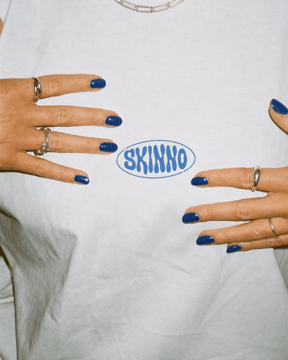 Close-up of hands with blue nail polish framing the Skinno logo on a white t-shirt