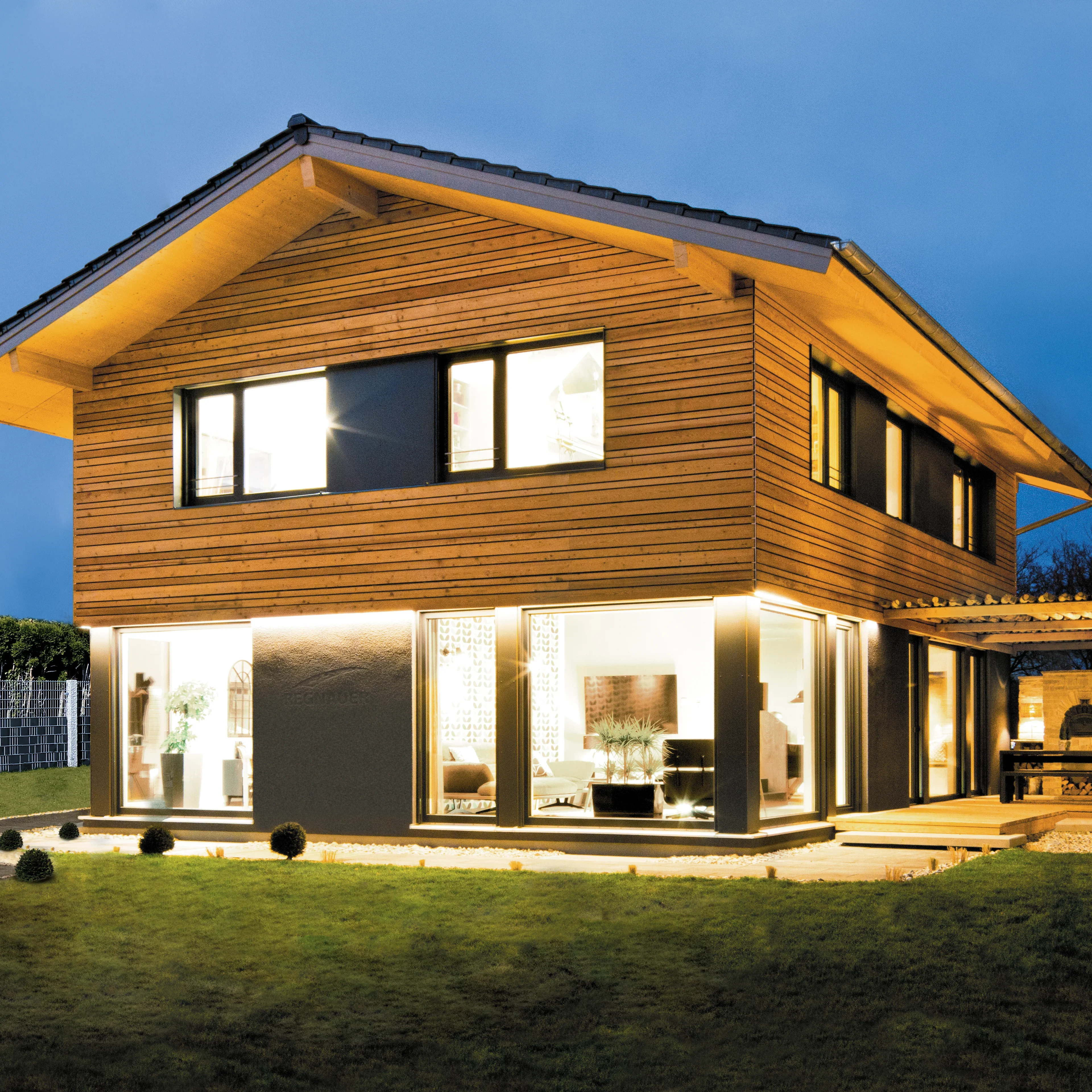 Vorderansicht eines zweigeschossigen Holzhauses mit großen Fenstern, die von warmem Licht erhellt werden, im Abendlicht.