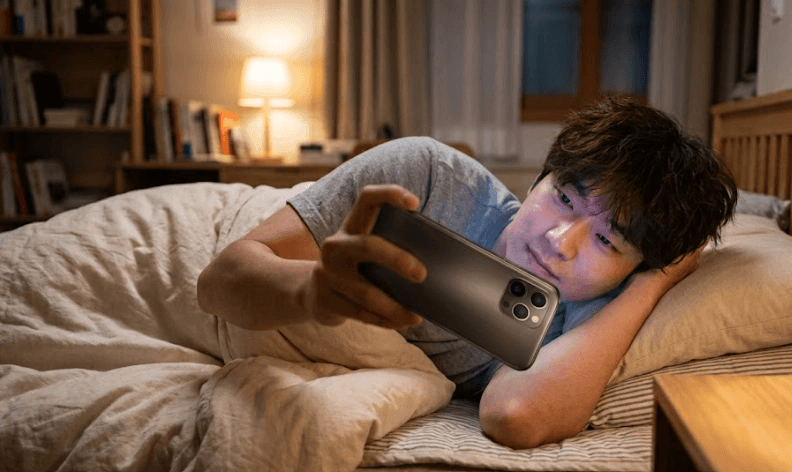 어두운 밤, 아늑한 침대 위에 옆으로 누워 스마트폰으로 영상을 시청하고 있는 젊은 남성의 모습입니다. 한 손으로 어두운 색상의 스마트폰을 가로로 쥐고 있으며, 화면에서 나오는 불빛이 남성의 얼굴을 밝히고 있습니다. 배경에는 은은한 스탠드 조명과 침실 가구들이 배치되어 편안한 야간 스마트폰 사용 환경을 보여줍니다.