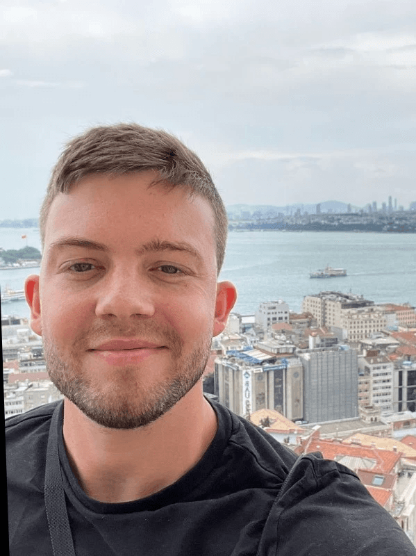 Hombre tomándose una selfie sobre un paisaje urbano costero con barcos y edificios lejanos.