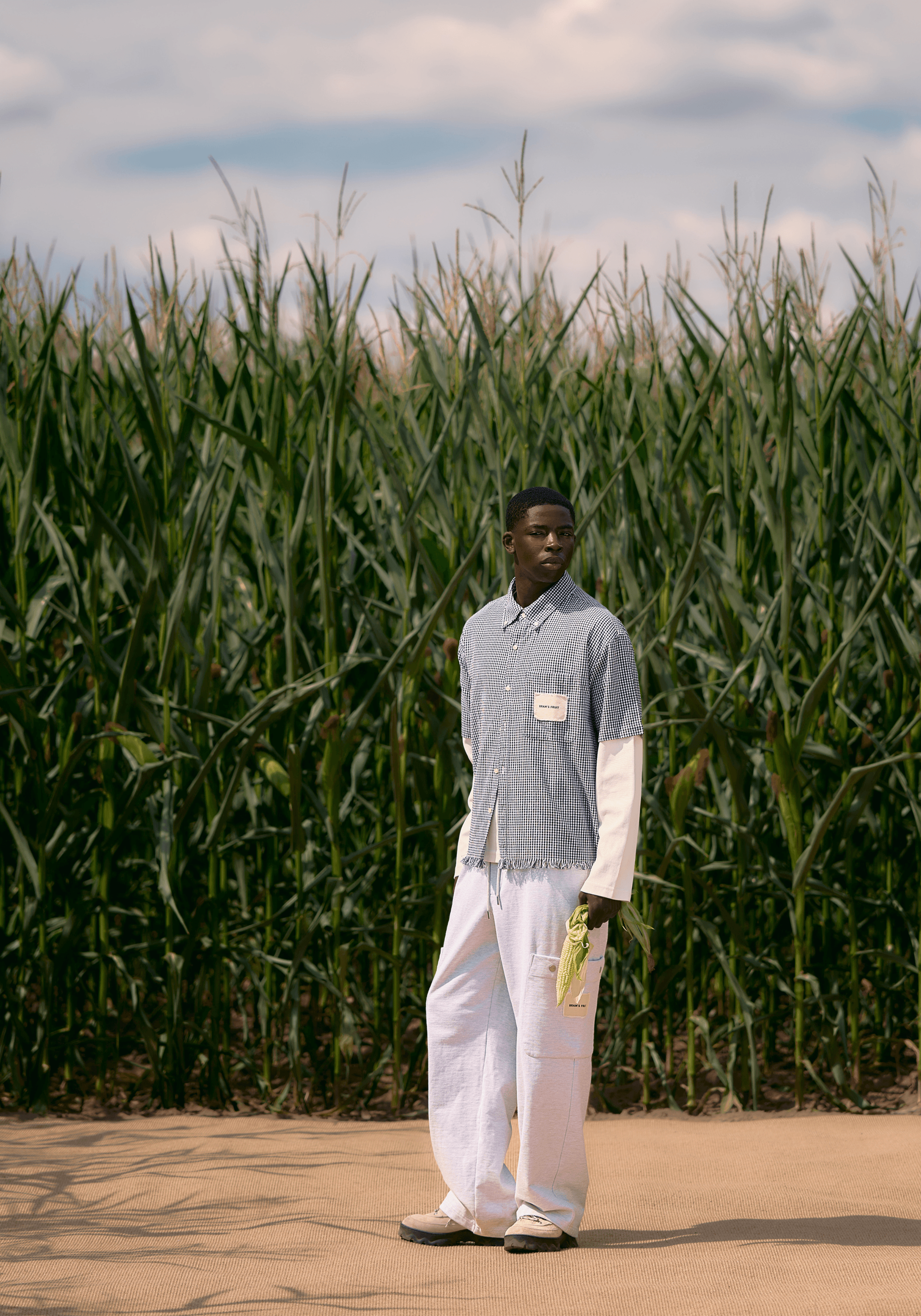 Devant un champ de mais, le mannequin porte un pantalonblanc et une chemise à petits carreaux effilochés de la marque Bram's Fruit