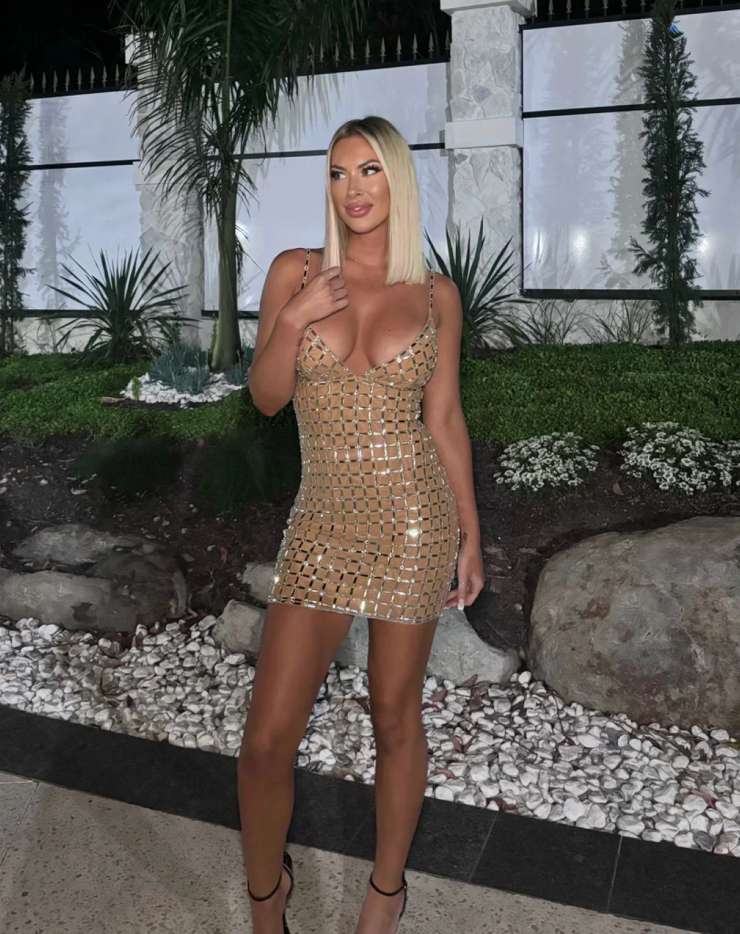 Topless waitress Vanessa posing in a gold mini dress in an outdoor garden setting for an Hourglass Gold Coast promotional photo.