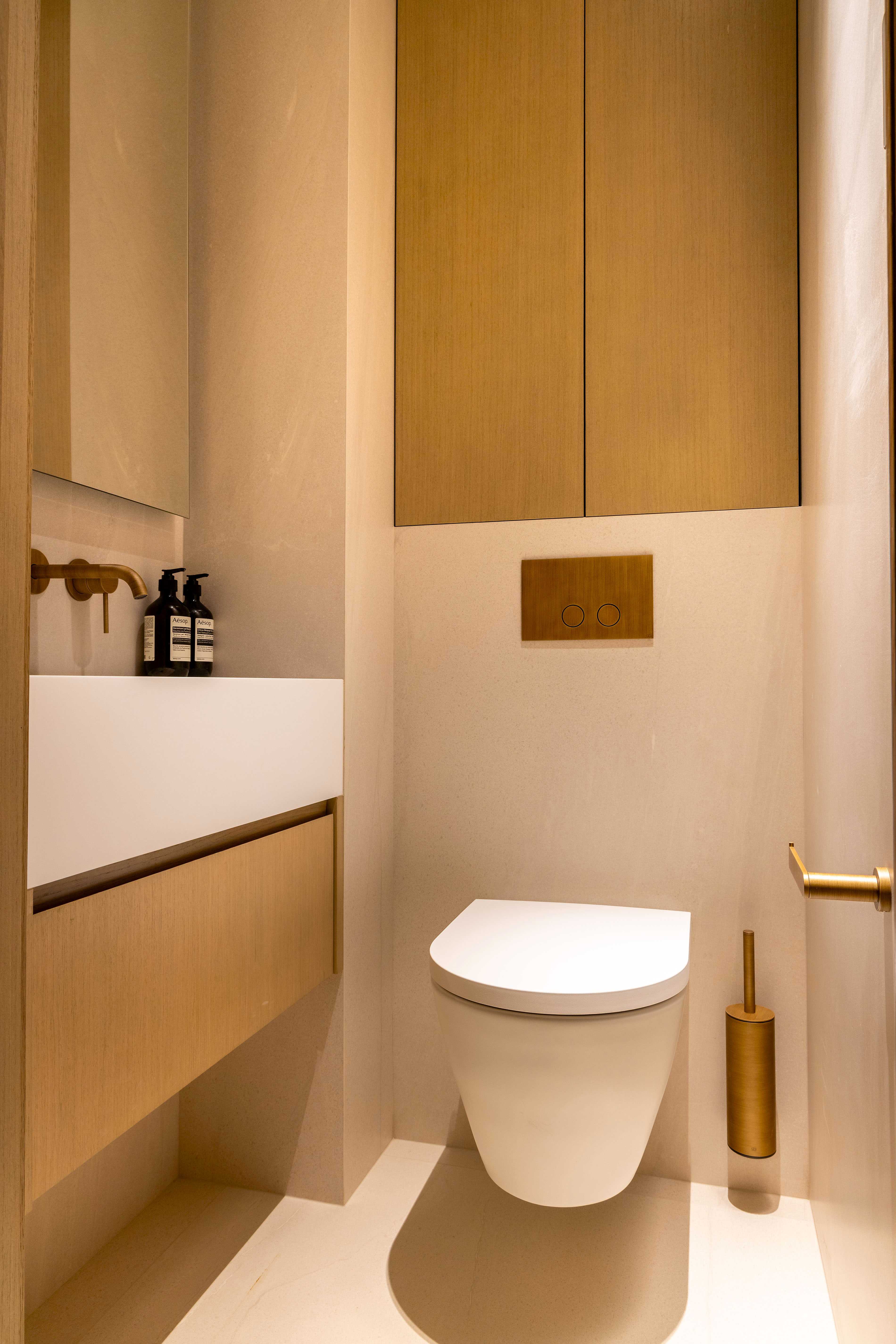 Toilettes suspendues avec plaque de commande dorée, rangement en bois clair et produits Aesop